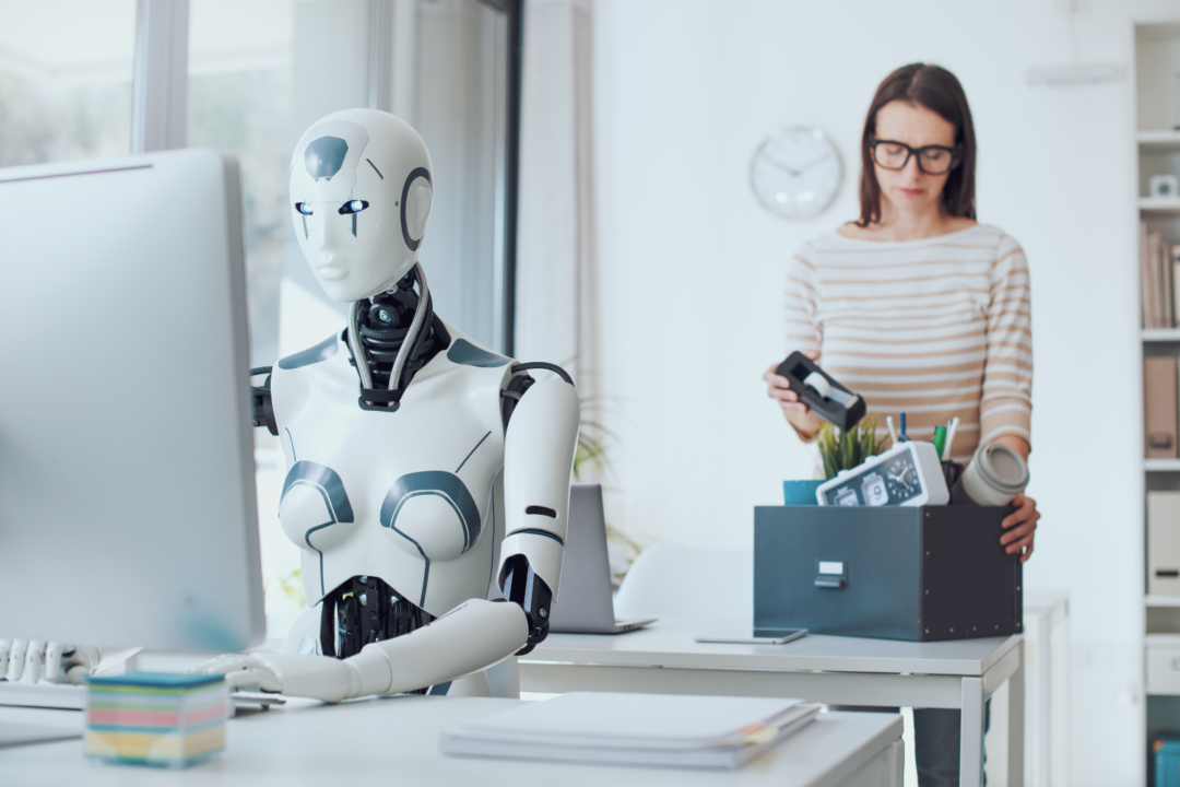 Humanoid robot working at an office desk as a woman packs her belongings, illustrating job displacement and the AI underclass