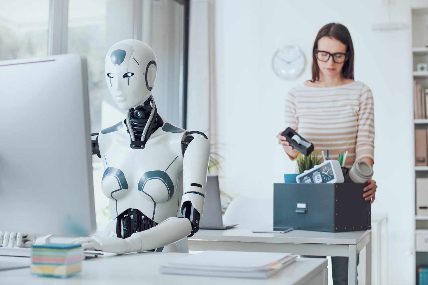Humanoid robot working at an office desk as a woman packs her belongings, illustrating job displacement and the AI underclass